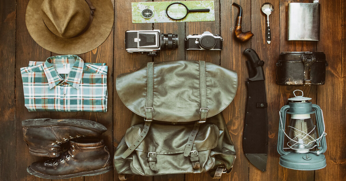 A collection of gear on a wooden background. There's a camera, knife, lantern, boots, a flask, and other various items.